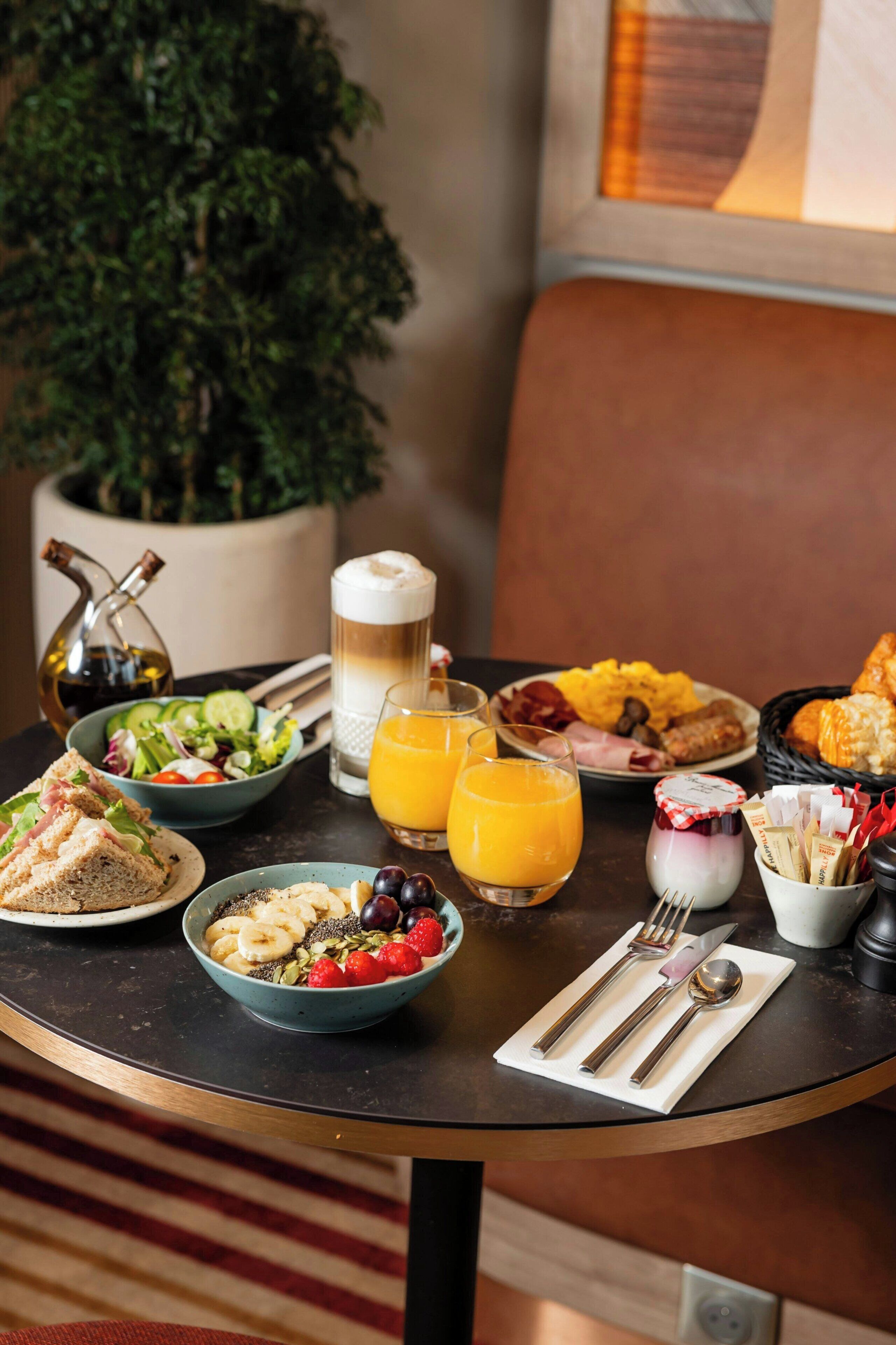 A breakfast table with various food items, a glass of orange juice, and a bottle of olive oil.