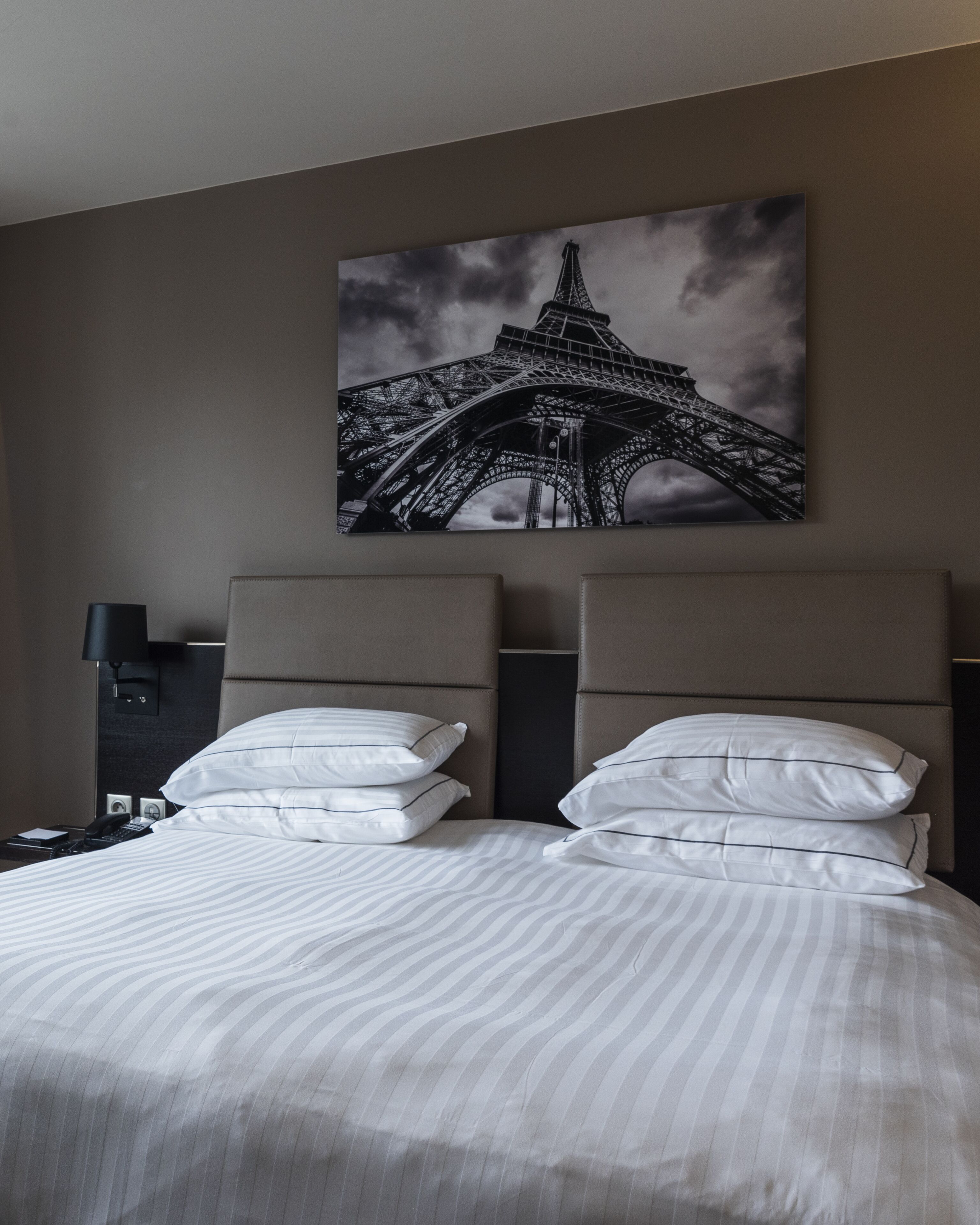 A hotel room with a bed, pillows, a nightstand, a lamp, and a large picture of the Eiffel Tower on the wall.