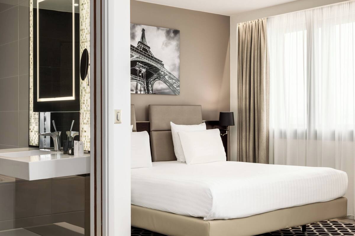 Ein modernes Hotelzimmer mit einem großen Bett, einem Nachttisch, einem Spiegel, einem Badezimmer mit Waschbecken und Blick auf den Eiffelturm durch das Fenster.