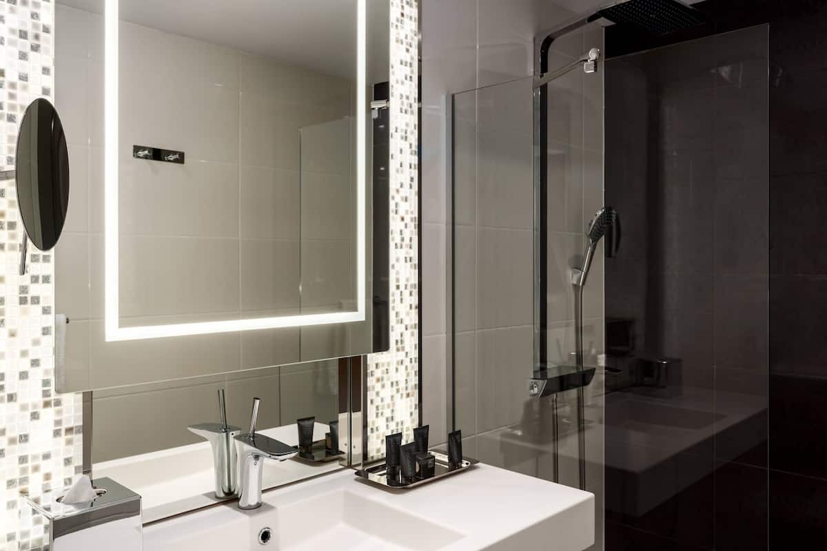 A modern bathroom with a large mirror, a glass shower enclosure, and a white sink with toiletries.