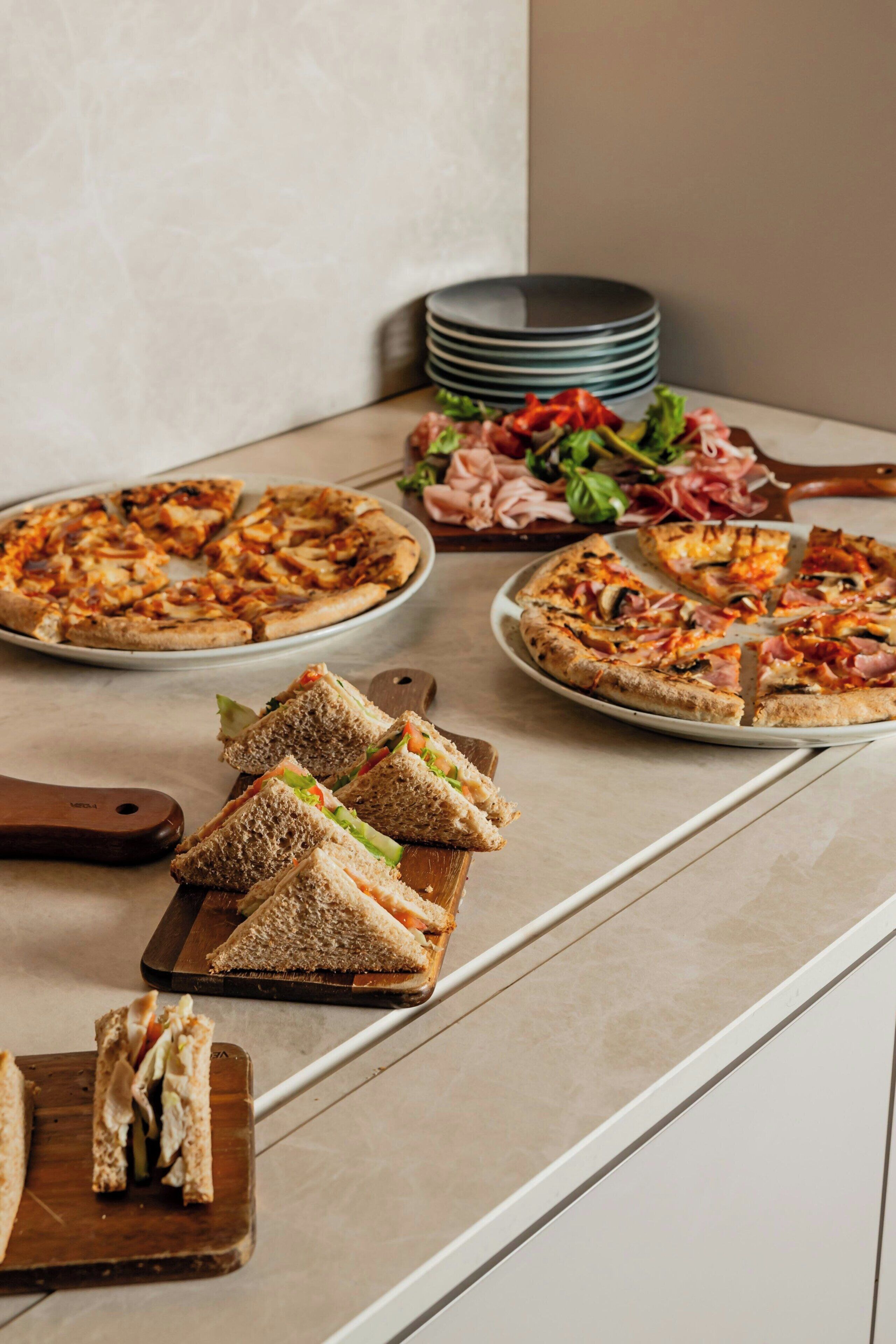 Eine Küchenarbeitsplatte mit zwei Pizzen, einem Sandwich und einem Stapel Teller.