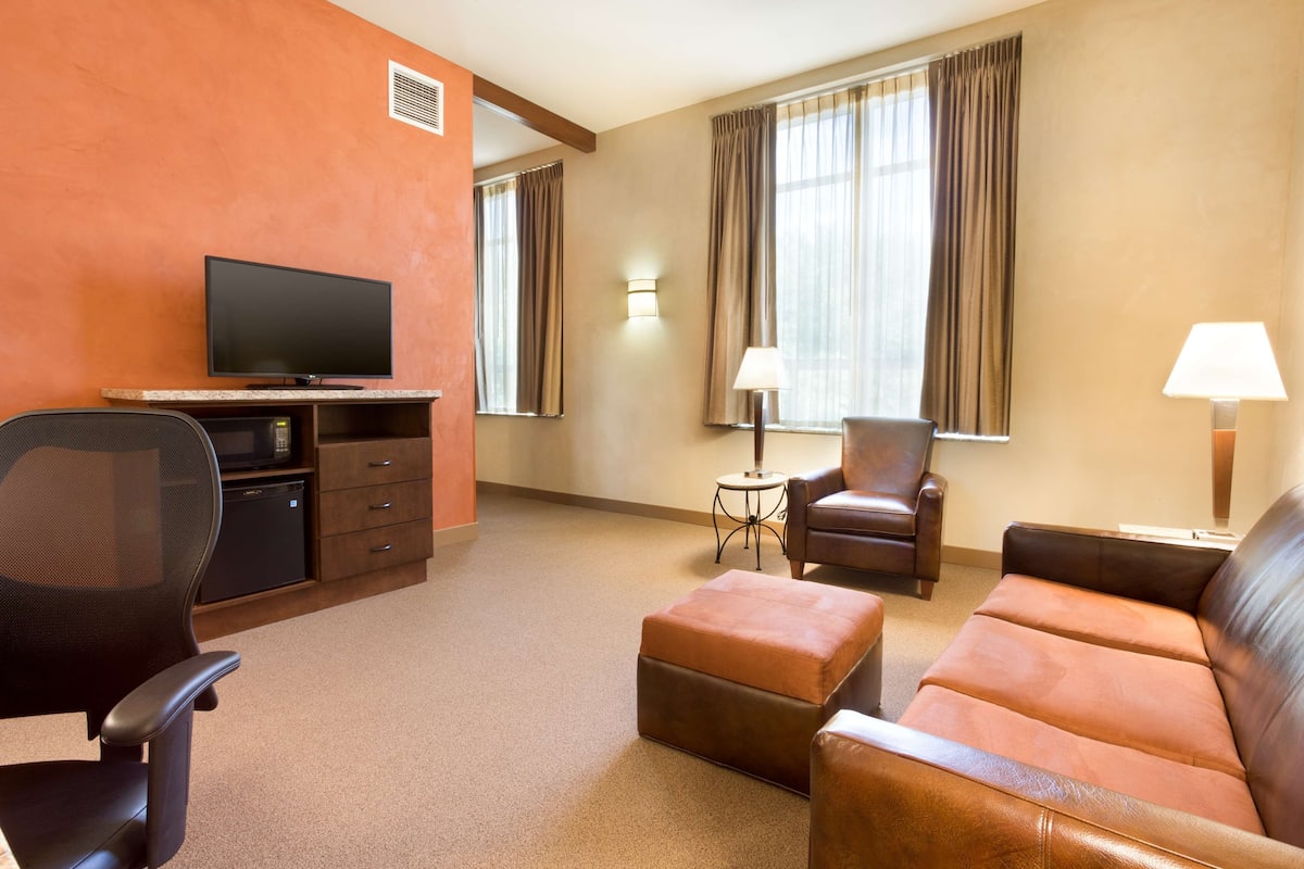 Une chambre d’hôtel équipée d’un téléviseur à écran plat, d’un bureau avec un ordinateur, d’un canapé en cuir marron, d’un fauteuil en cuir marron, d’une petite table ronde avec une lampe et d’une fenêtre avec des rideaux.