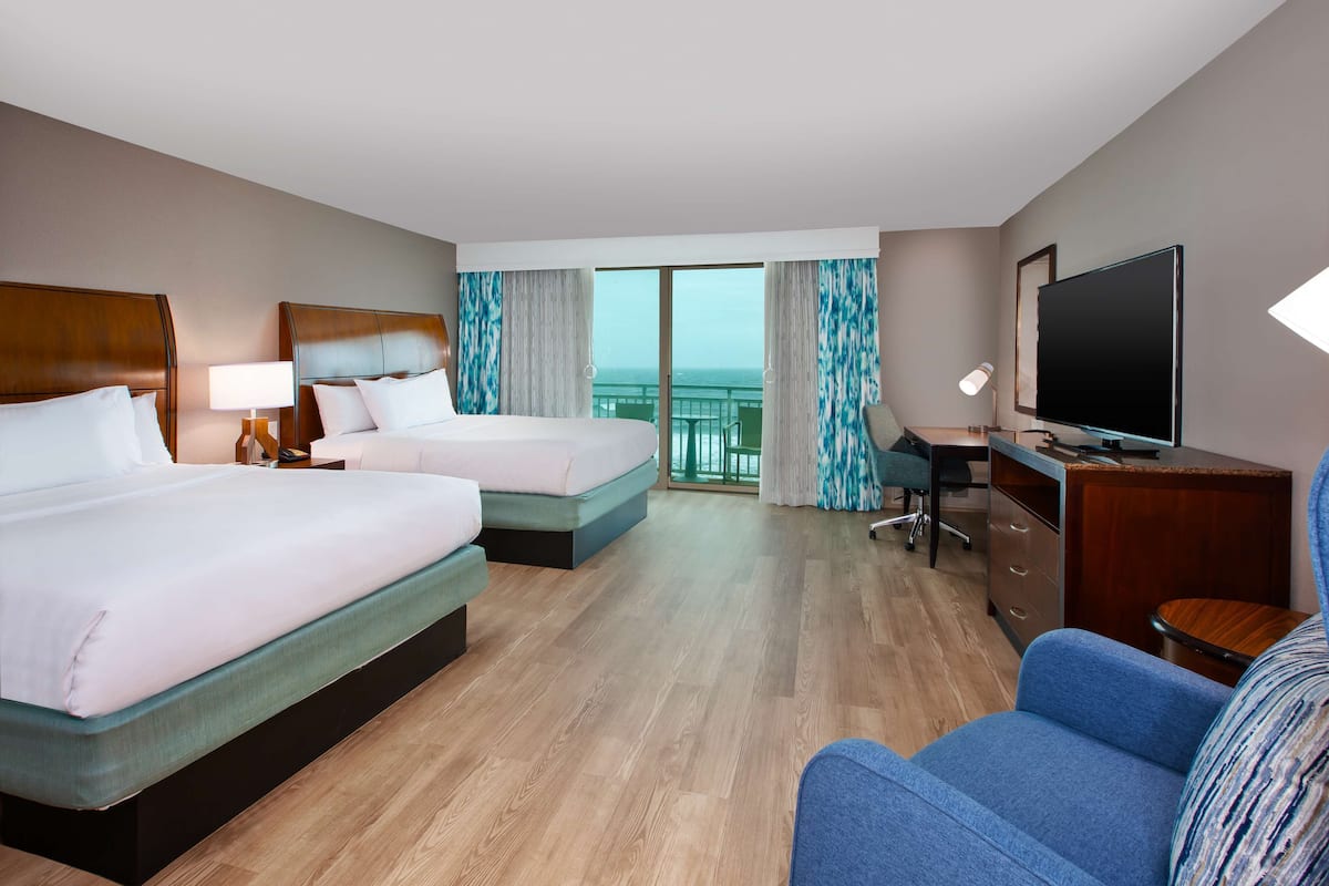 A hotel room with two beds, a desk, a television, and a view of the ocean.