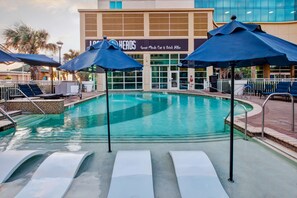 A swimming pool with lounge chairs and umbrellas, a building with a sign reading "Great Minds Eat & Drink Alike," and a palm tree in the background. - Hilton Garden Inn Virginia Beach Oceanfront (Virginia Beach)