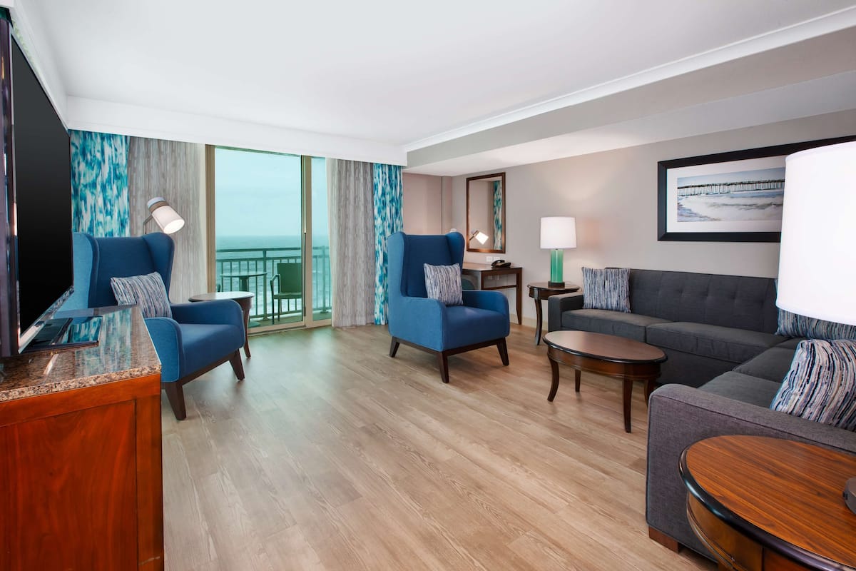 A hotel room with a wooden floor, two blue armchairs, a grey sofa, a small round table, a TV, a lamp, a framed picture, and a view of the sea through large windows.