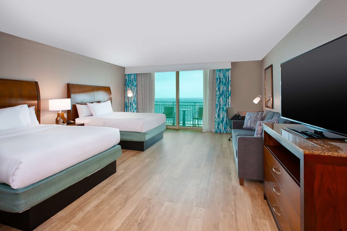 A hotel room with two beds, a TV, a desk, and a balcony with a view.