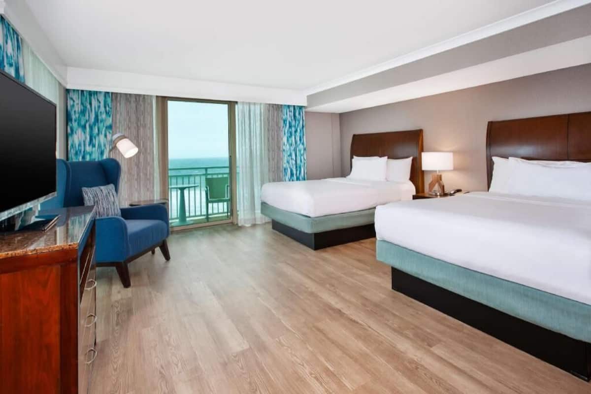 A hotel room with two beds, a desk, a TV, and a balcony with curtains.