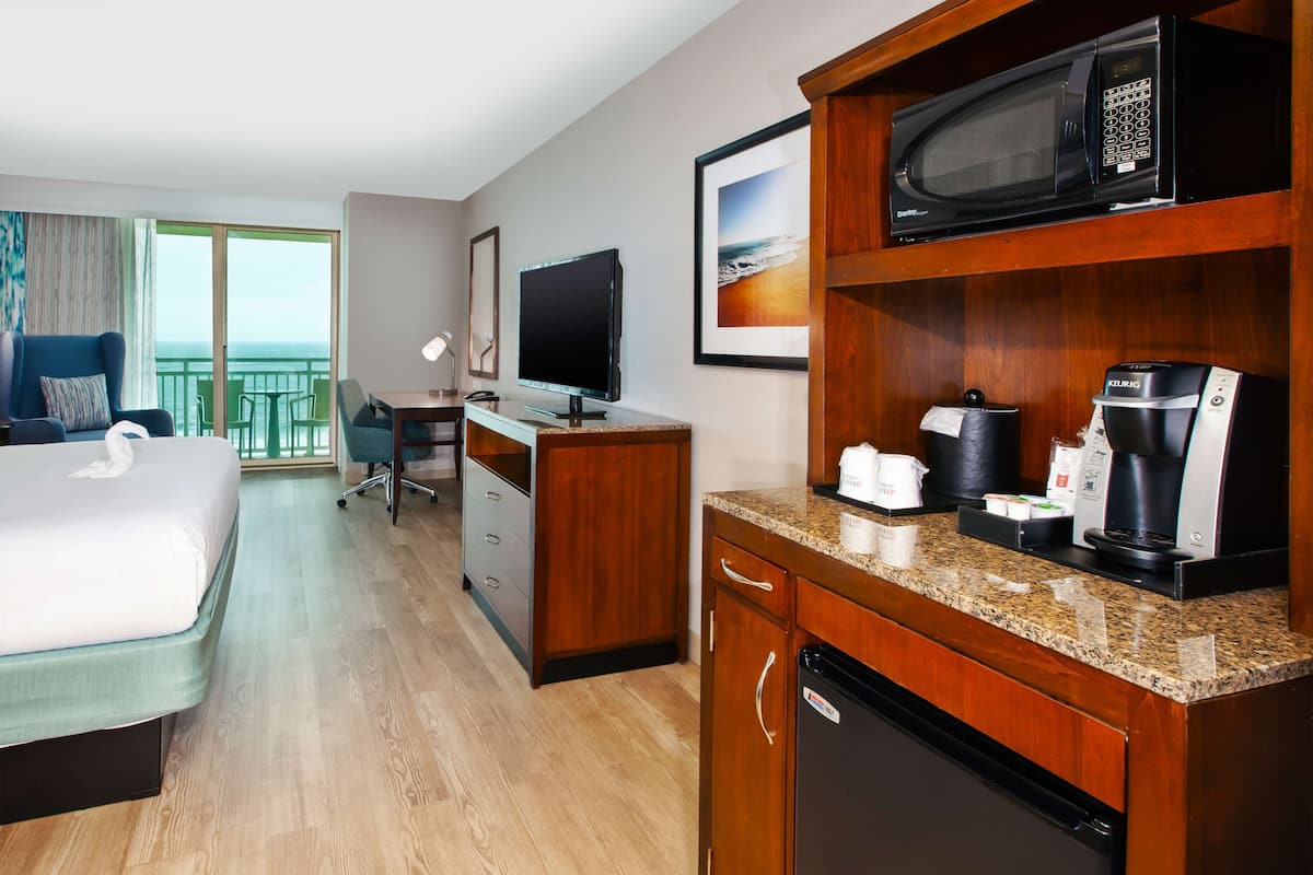 A hotel room with a bed, a desk with a computer, a microwave, a coffee maker, and a view of the ocean.