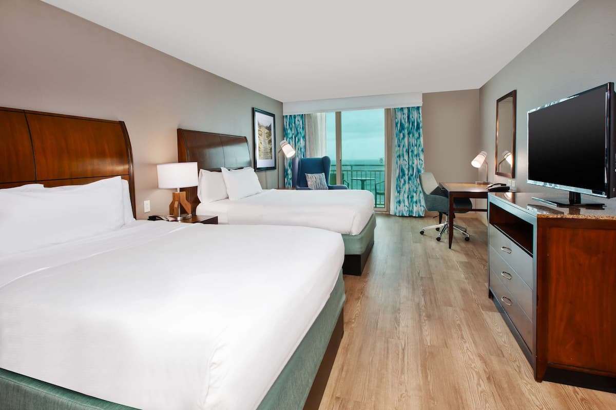 A hotel room with two beds, a desk, a TV, and a balcony with a view.