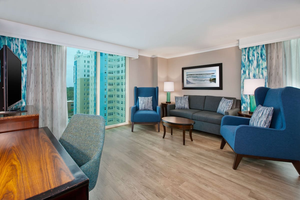 A modern hotel room with a wooden floor, a large window with a city view, a sofa, two armchairs, a coffee table, and a TV.