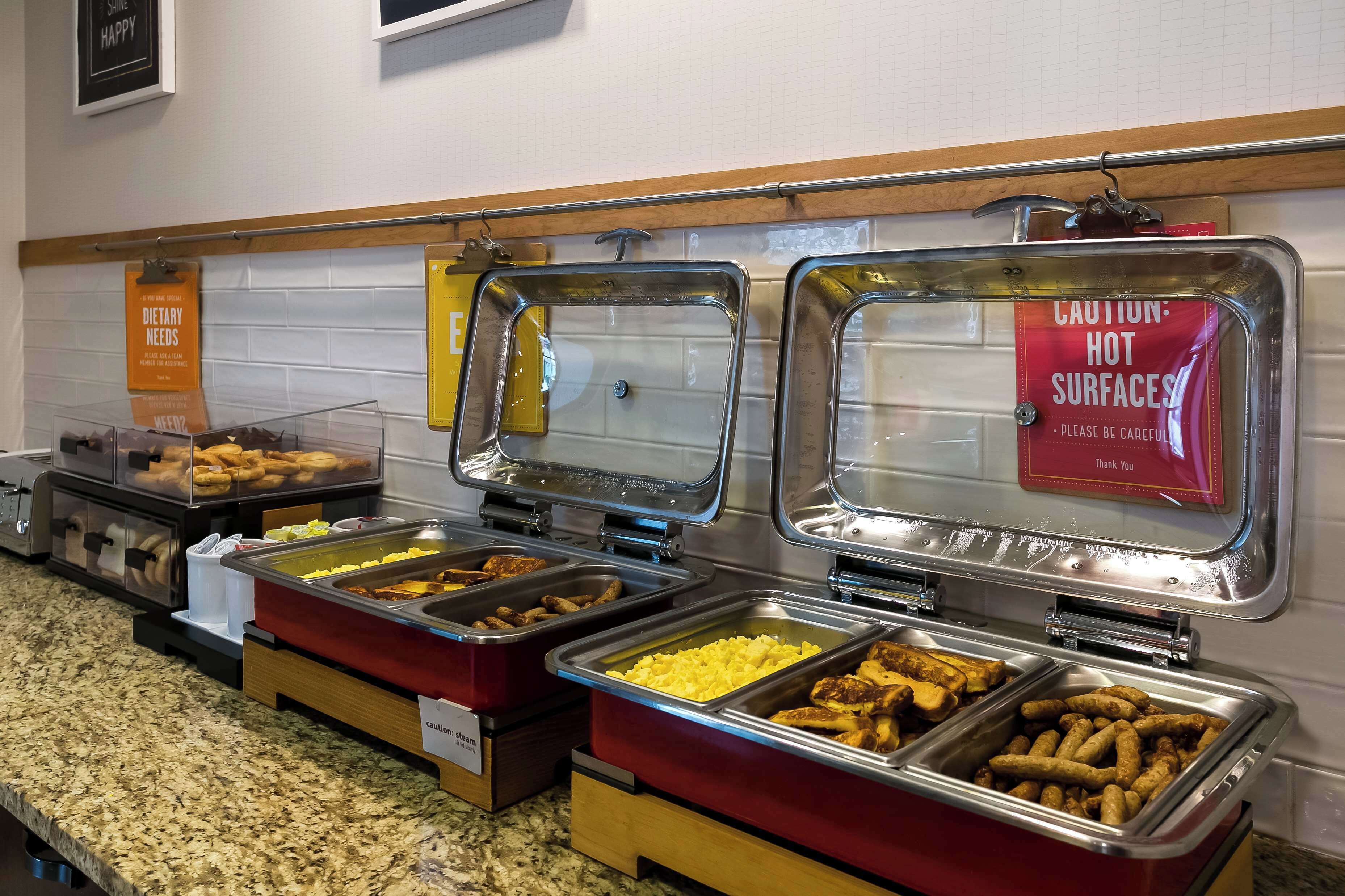 Un buffet proposant divers plats présentés dans des récipients en métal, un panneau d’avertissement indiquant que certaines surfaces sont chaudes, et un panneau indiquant les besoins alimentaires spécifiques.