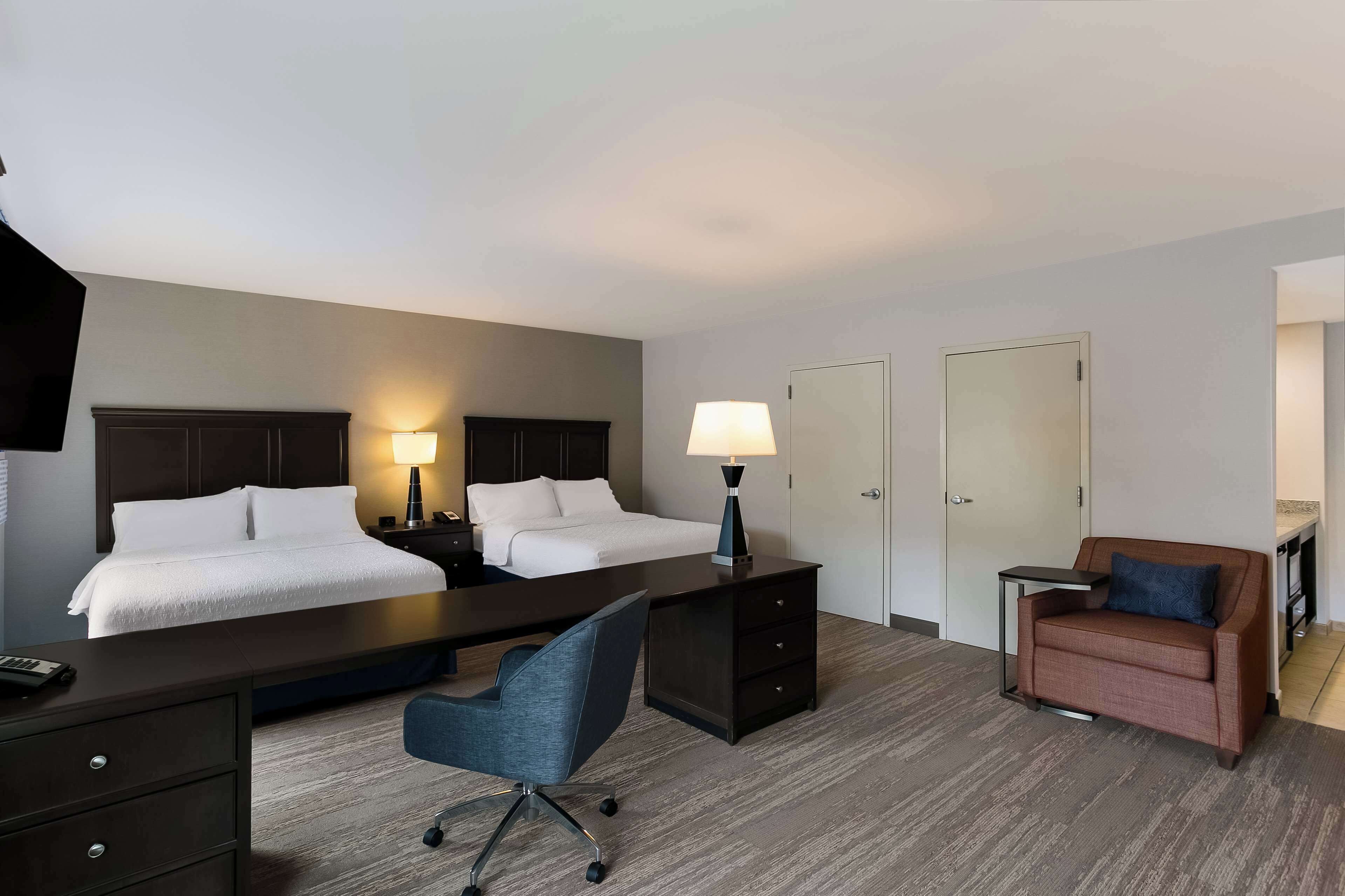 A hotel room with two beds, a desk, a chair, a lamp, a sofa, and a wardrobe.