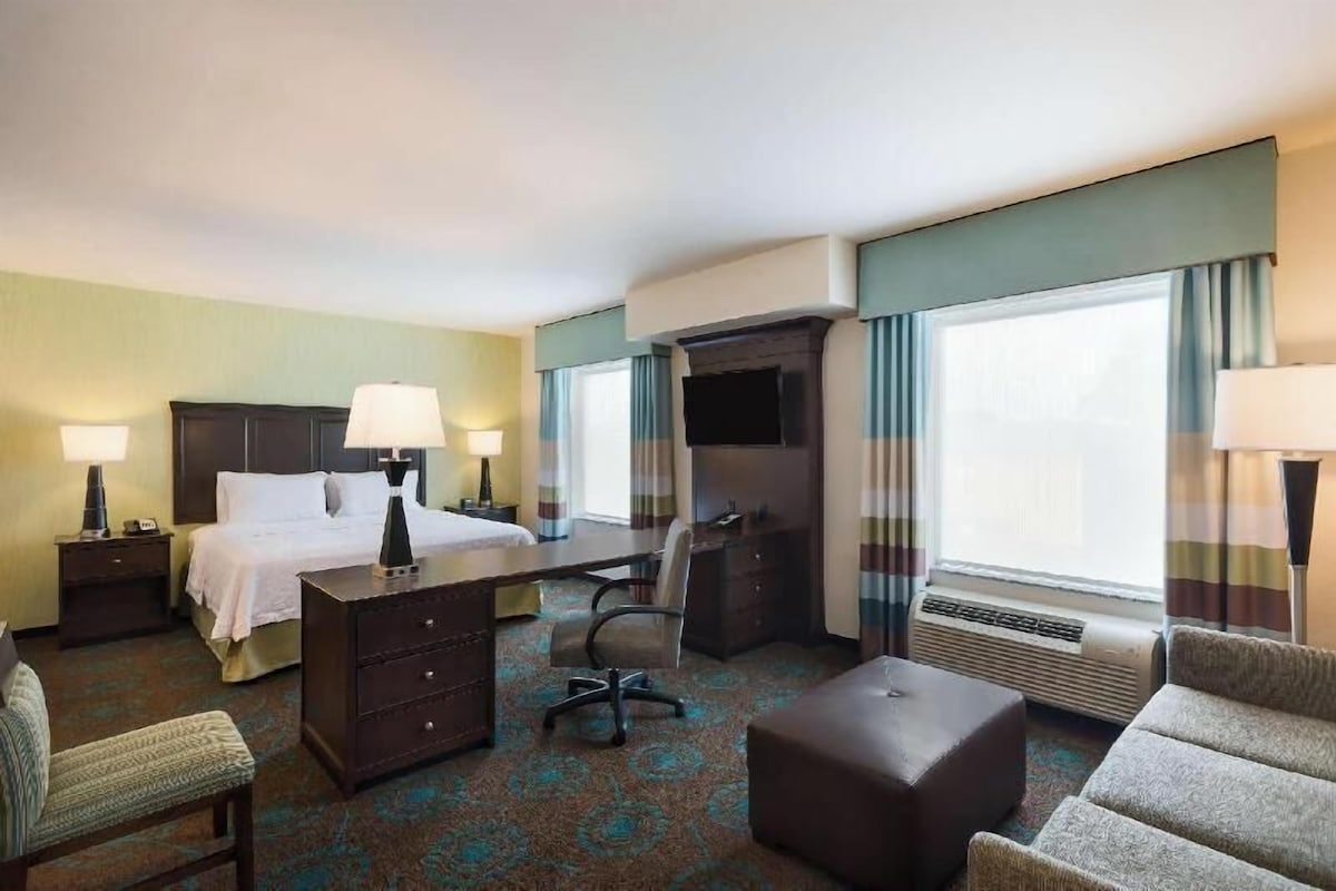 A hotel room with a bed, desk, chair, ottoman, and a large window.