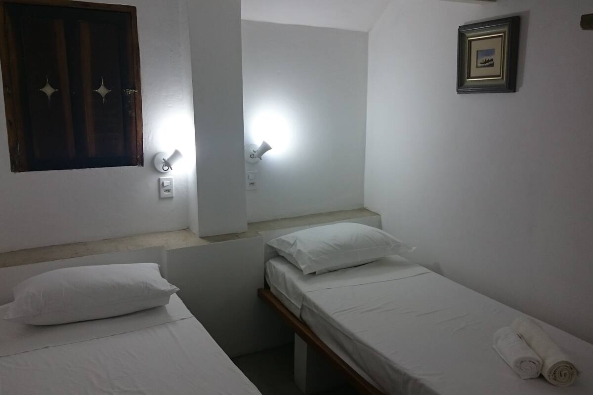 A small, clean room with two single beds, a wooden door, a framed picture on the wall, and a window with curtains.