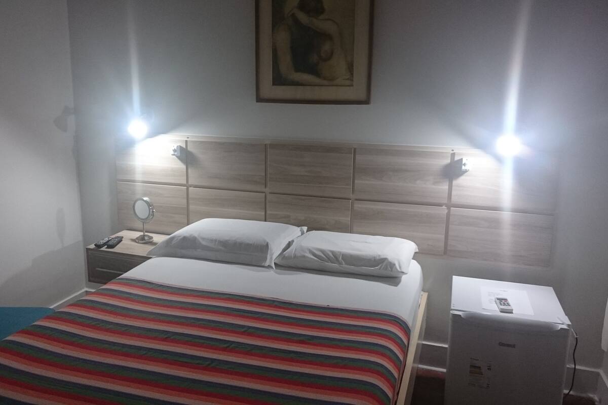 A hotel room with a bed, a nightstand with a mirror, a blue chair, and a heater.