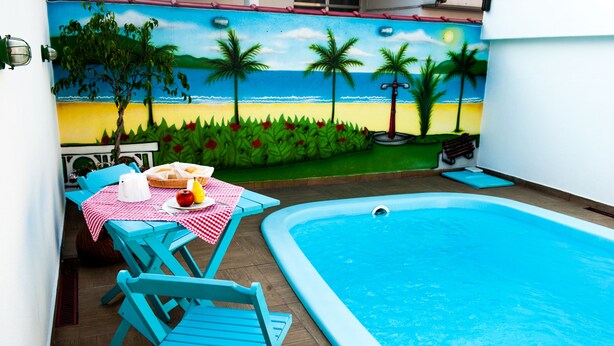 Zona de piscina con un mural que representa una playa, una mesa con mantel a cuadros y sillas azules.