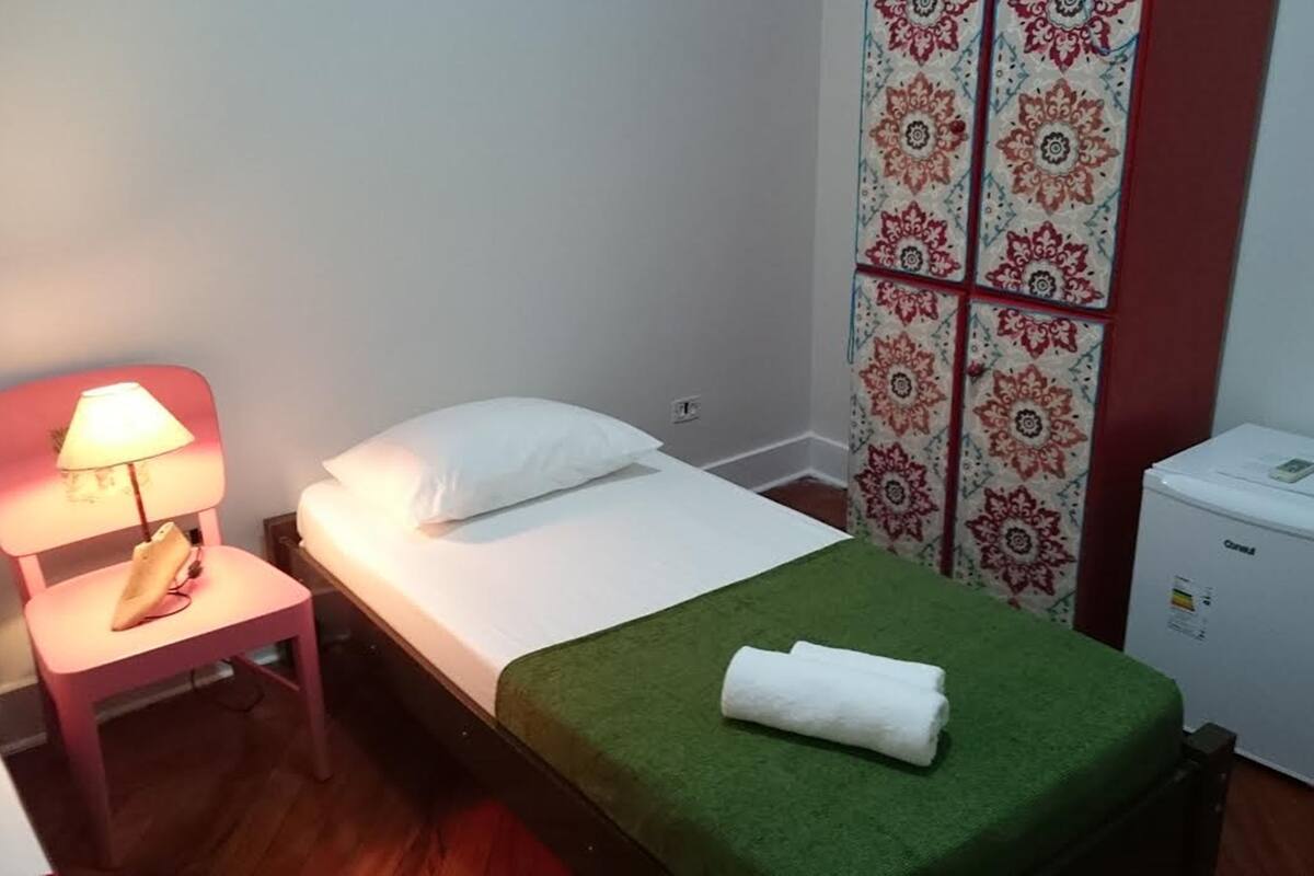 A small, neatly arranged bedroom with a single bed, a bedside table with a lamp, a small refrigerator, and patterned curtains.