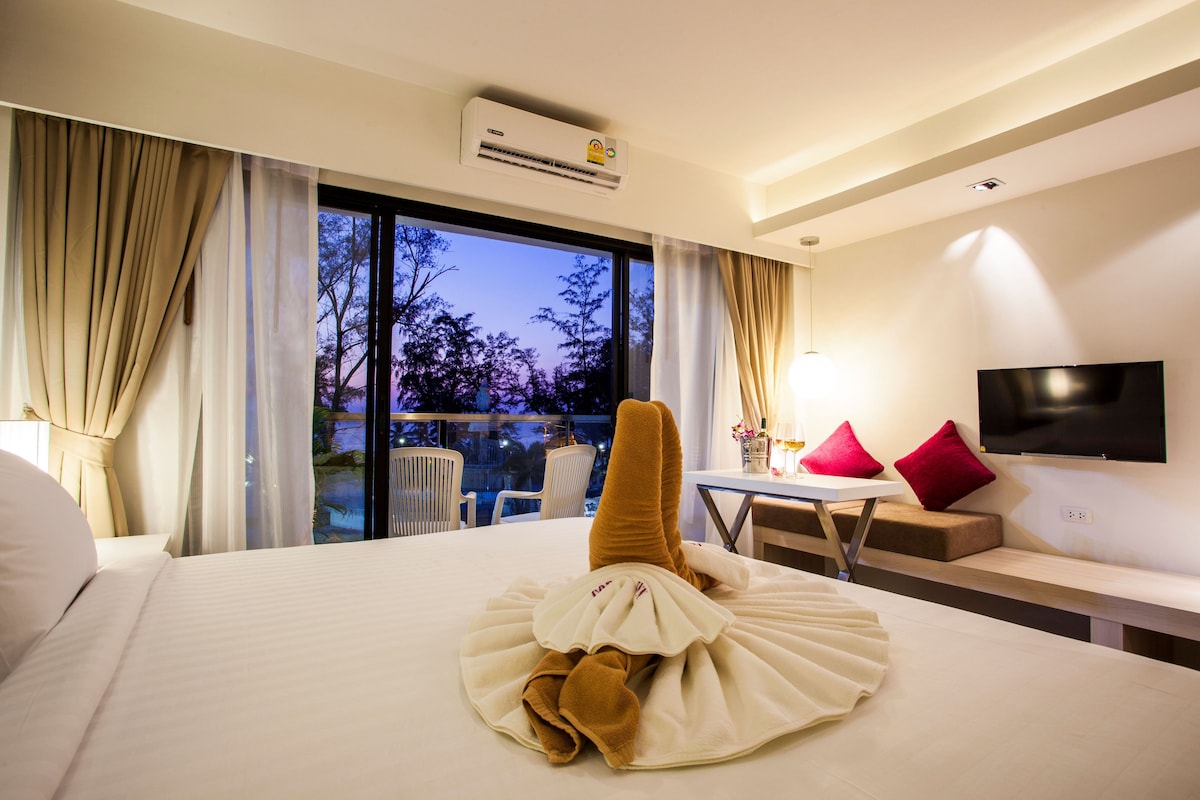 A hotel room with a large bed, a flat-screen TV, a dining table, and a view of trees through the window.
