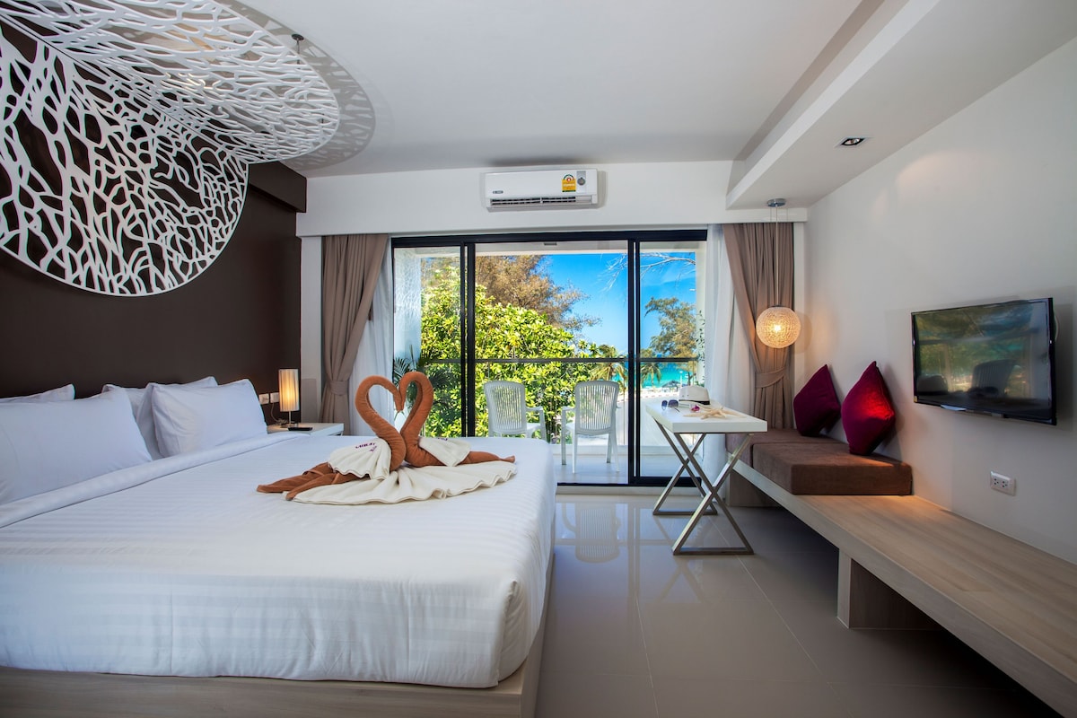 A hotel room with a large bed, a desk with a chair, a TV, and a balcony with a view of trees and sky.
