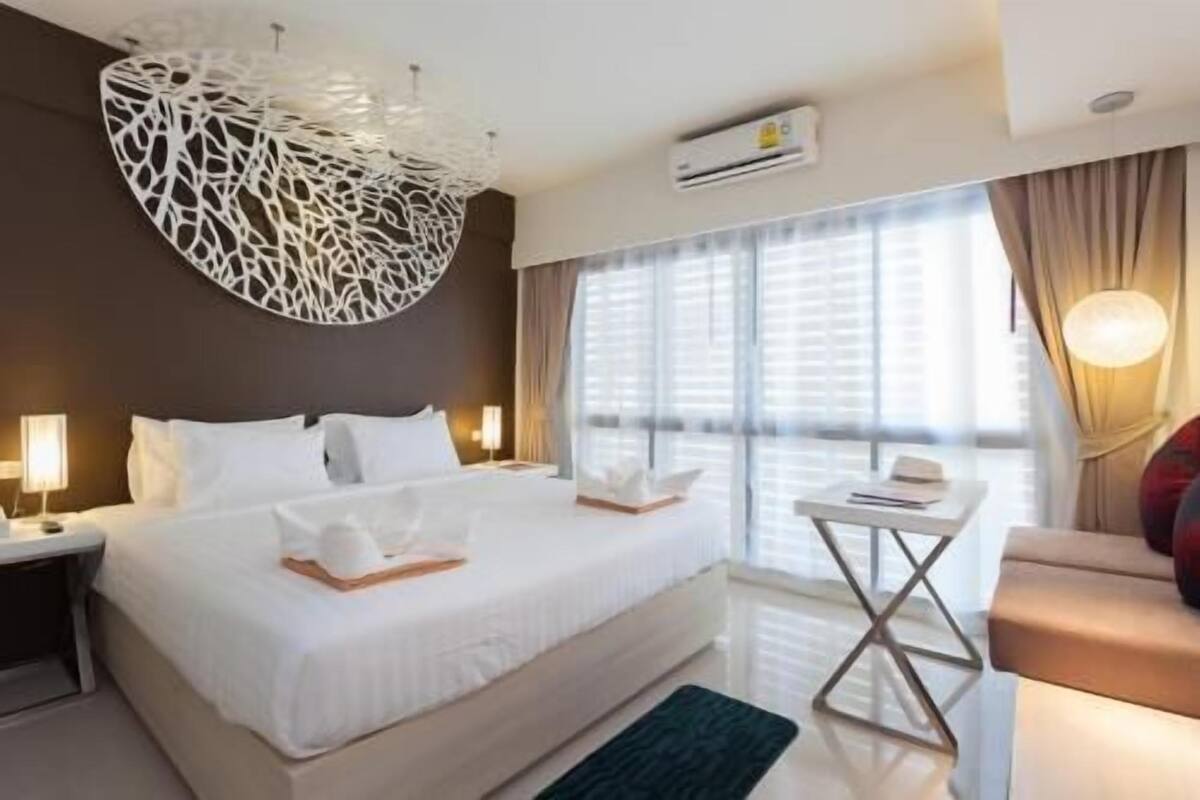 Una habitación de hotel moderna con una cama grande, lámparas de noche, una mesita y vistas al exterior a través de amplios ventanales.