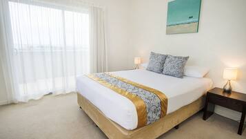 A neatly made bed with a gold and white bedspread, two bedside lamps, a nightstand, and a painting of a beach scene on the wall.