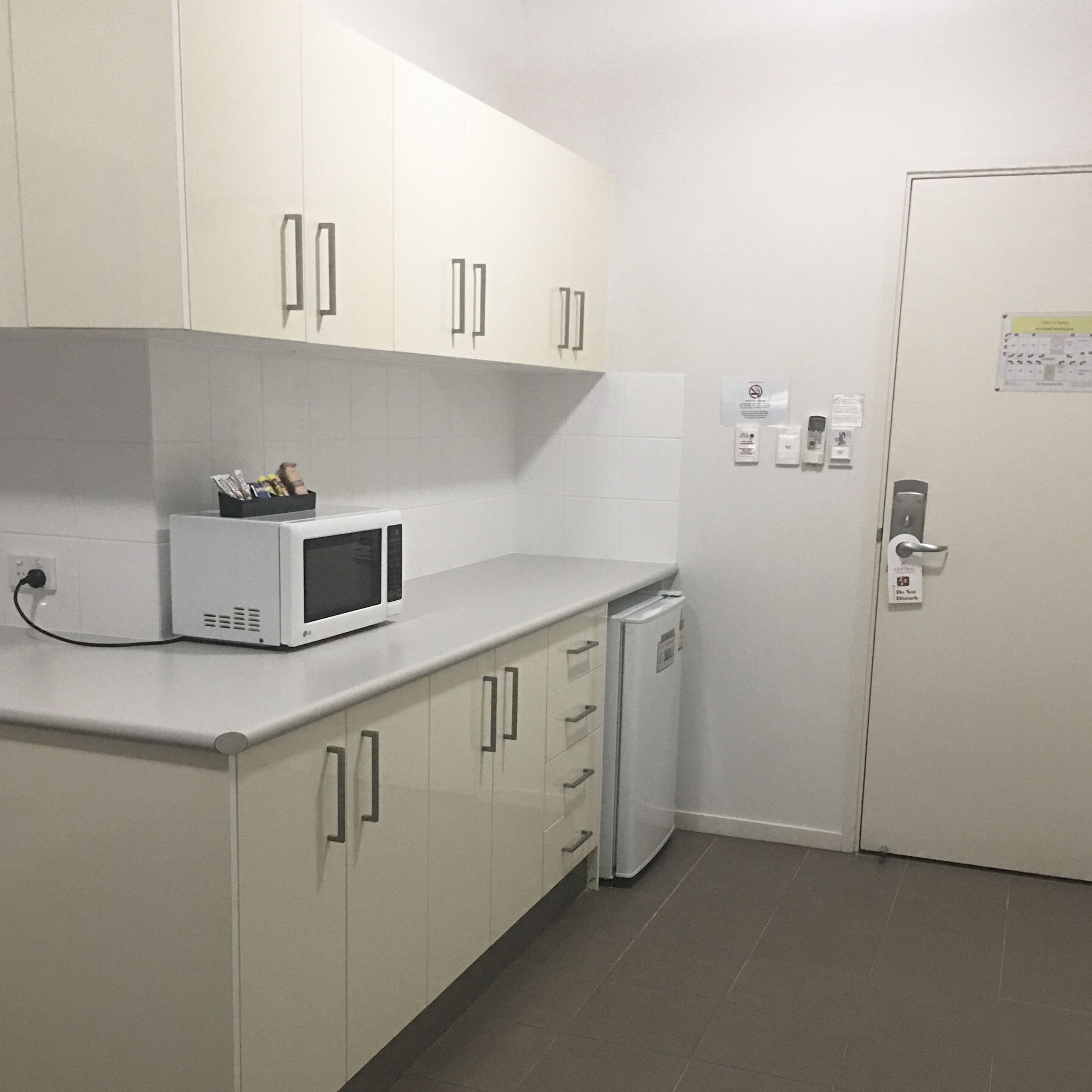 Uma pequena cozinha compacta com armários brancos, micro-ondas e geladeira. Há tomadas elétricas e uma porta com tranca.