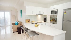 A modern kitchen with white cabinets, a central island with stools, a coffee machine, and a microwave. - Gladstone City Central Apartment Hotel (Gladstone)