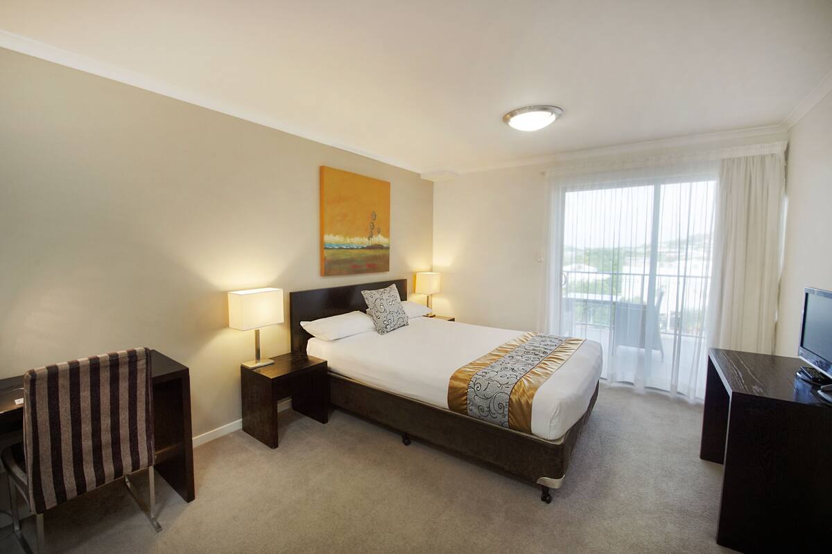 A hotel room with a large bed, two bedside tables with lamps, a desk with a TV, and a window with curtains.