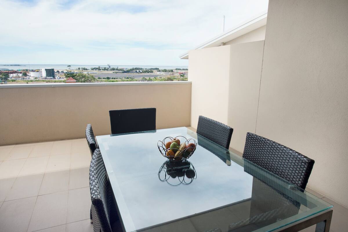 A glass-top table with a fruit bowl, wicker chairs, and a view of the cityscape.