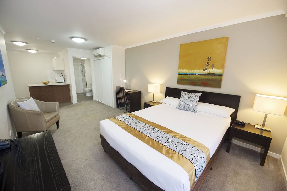 A hotel room with a large bed, a desk, a chair, a desk lamp, a bedside table, and a painting on the wall.
