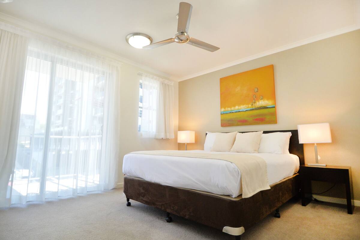 A hotel room with a large bed, two bedside lamps, a ceiling fan, and a painting on the wall.