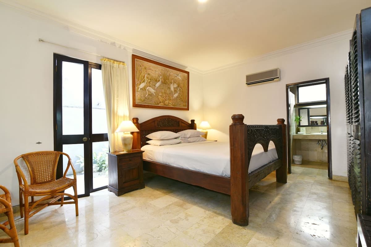 A bedroom with a wooden bed, two bedside tables with lamps, a wicker chair, a large window with curtains, and a painting on the wall.