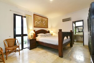 Un dormitorio con cama de madera, dos mesitas de noche con lámparas, una silla de mimbre, un cuadro grande y un baño visible a través de una puerta abierta.