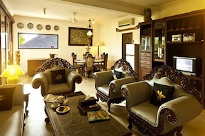 A living room with a sofa, armchairs, a coffee table, and a television. There is a dining table with chairs and a chandelier.