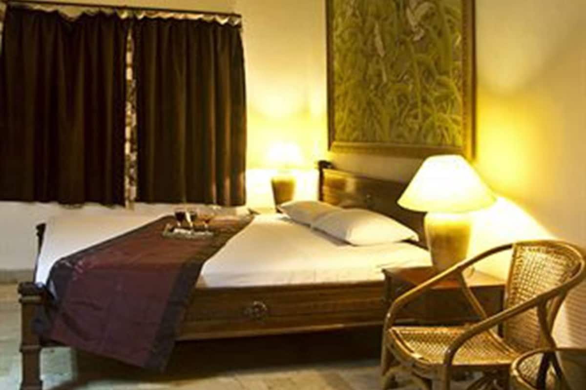 A hotel room with a bed, a ceiling fan, a nightstand with a lamp, a chair, and a large painting on the wall.
