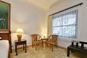 Une pièce comprenant un lit, deux fauteuils en osier, une petite table avec une lampe, un banc et une fenêtre ornée de rideaux légers.