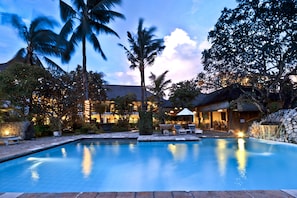 A swimming pool with lounge chairs and umbrellas, surrounded by palm trees and a building with outdoor seating.