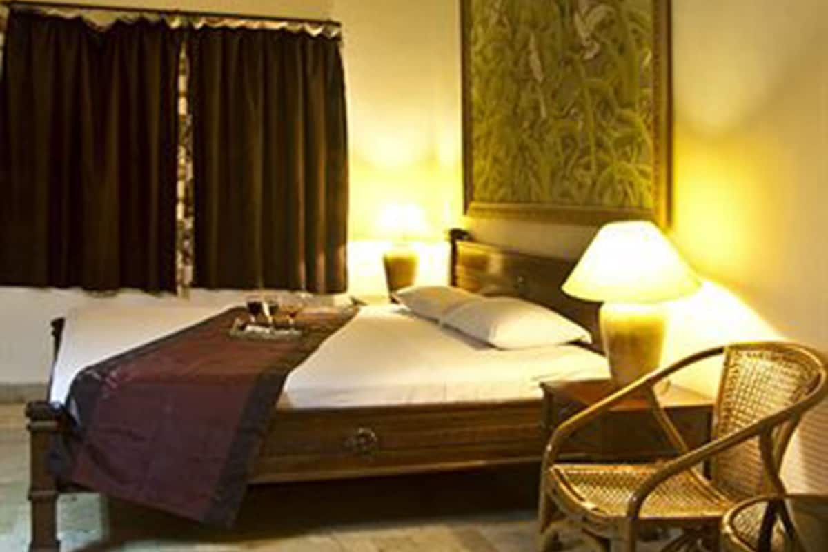 A hotel room with a bed, a ceiling fan, a nightstand with a lamp, a chair, and a painting on the wall.
