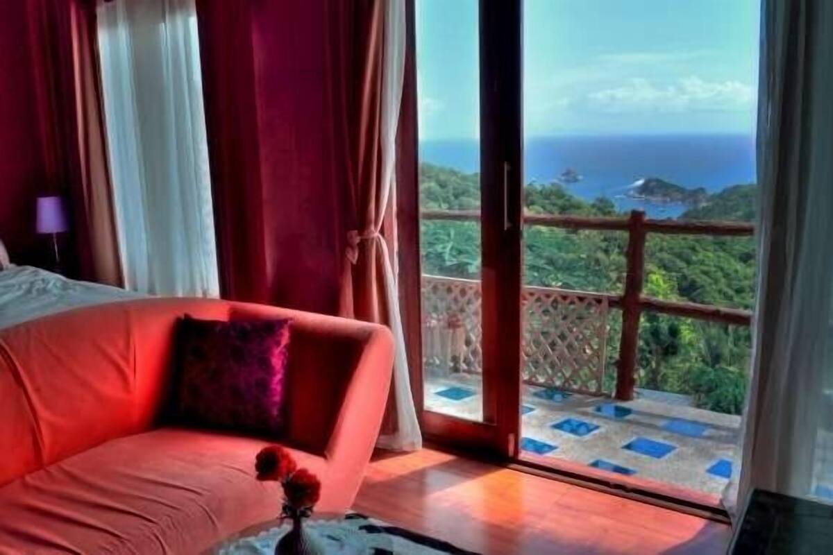 A room with a red sofa, a view of the ocean, and a balcony with a wooden railing.