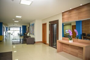 A hotel lobby with a reception desk, a large painting, an elevator, and a seating area with chairs and a potted plant. - Hit Hotel (Salvador)