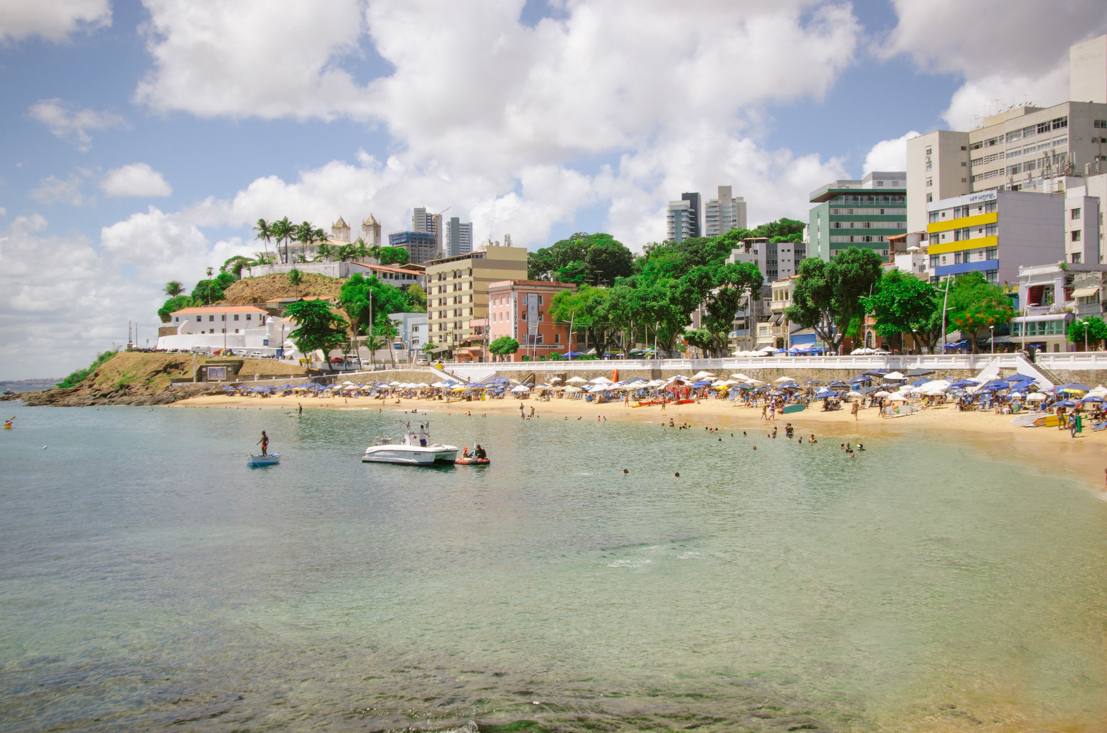 A beach with clear water, a boat, and a cityscape with buildings and palm trees in the background.
