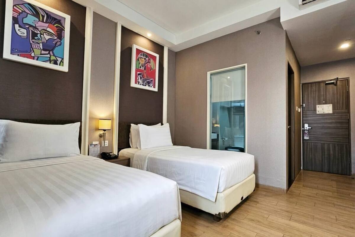 A hotel room with two beds, a bedside table with a lamp, a large window, and a door.