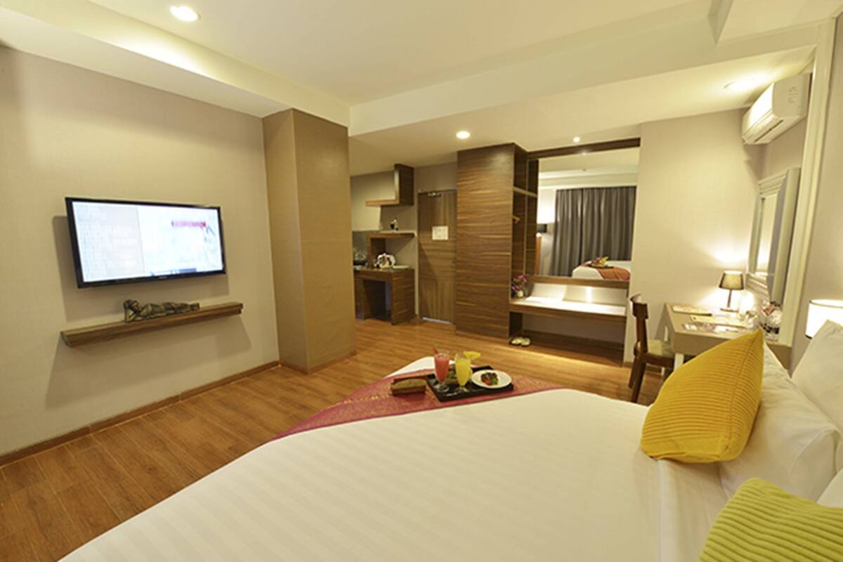 A hotel room with a large bed, a TV mounted on the wall, a small kitchenette, and a bathroom with a mirror.