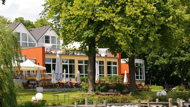 A lakeside hotel with outdoor seating, a dining area, and a view of the water. - DämeritzSeehotel (Berlin)