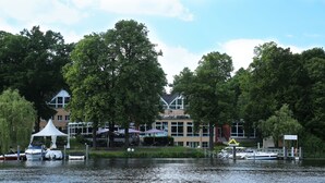 A lakeside view with boats docked, a white tent, and a building with large windows and a sloped roof. - DämeritzSeehotel (Berlin)