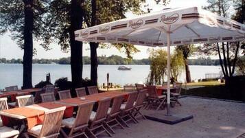 An outdoor patio with wooden tables and chairs, a large umbrella, and a view of a lake with a boat in the background.