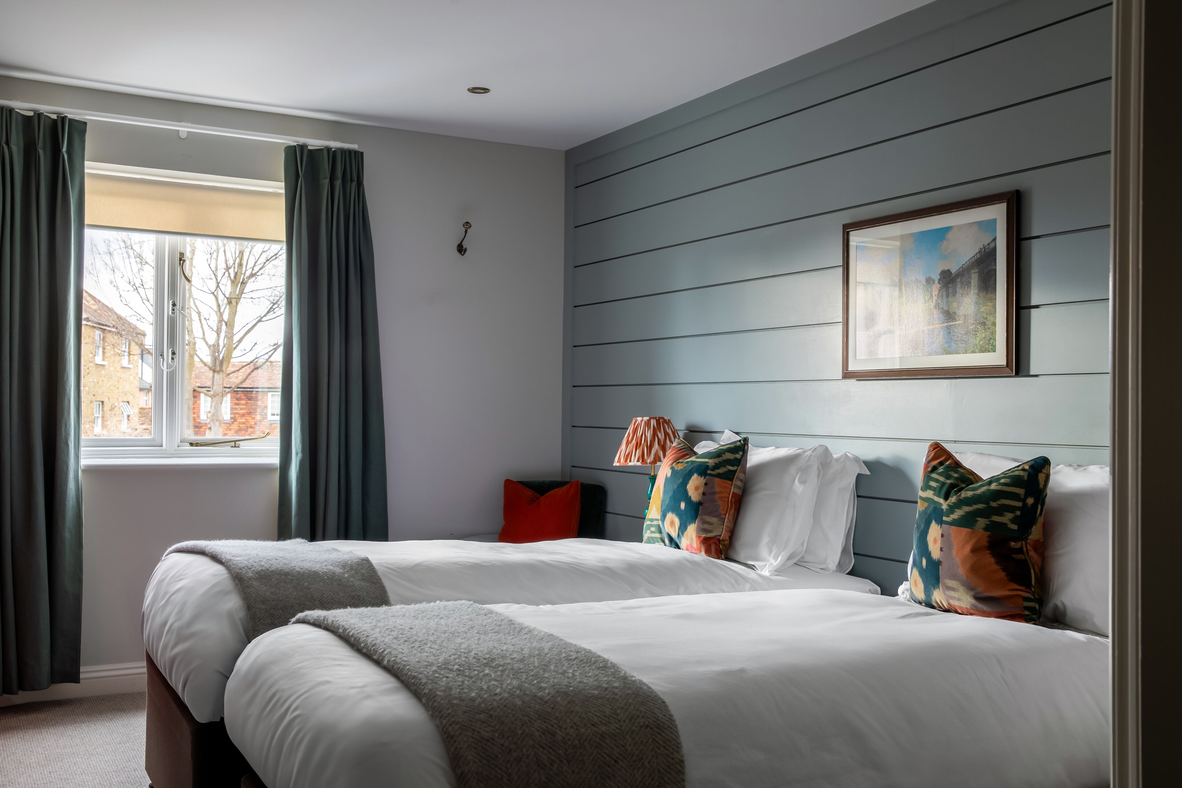 A hotel room with two beds, a window with curtains, a painting on the wall, and a red pillow.