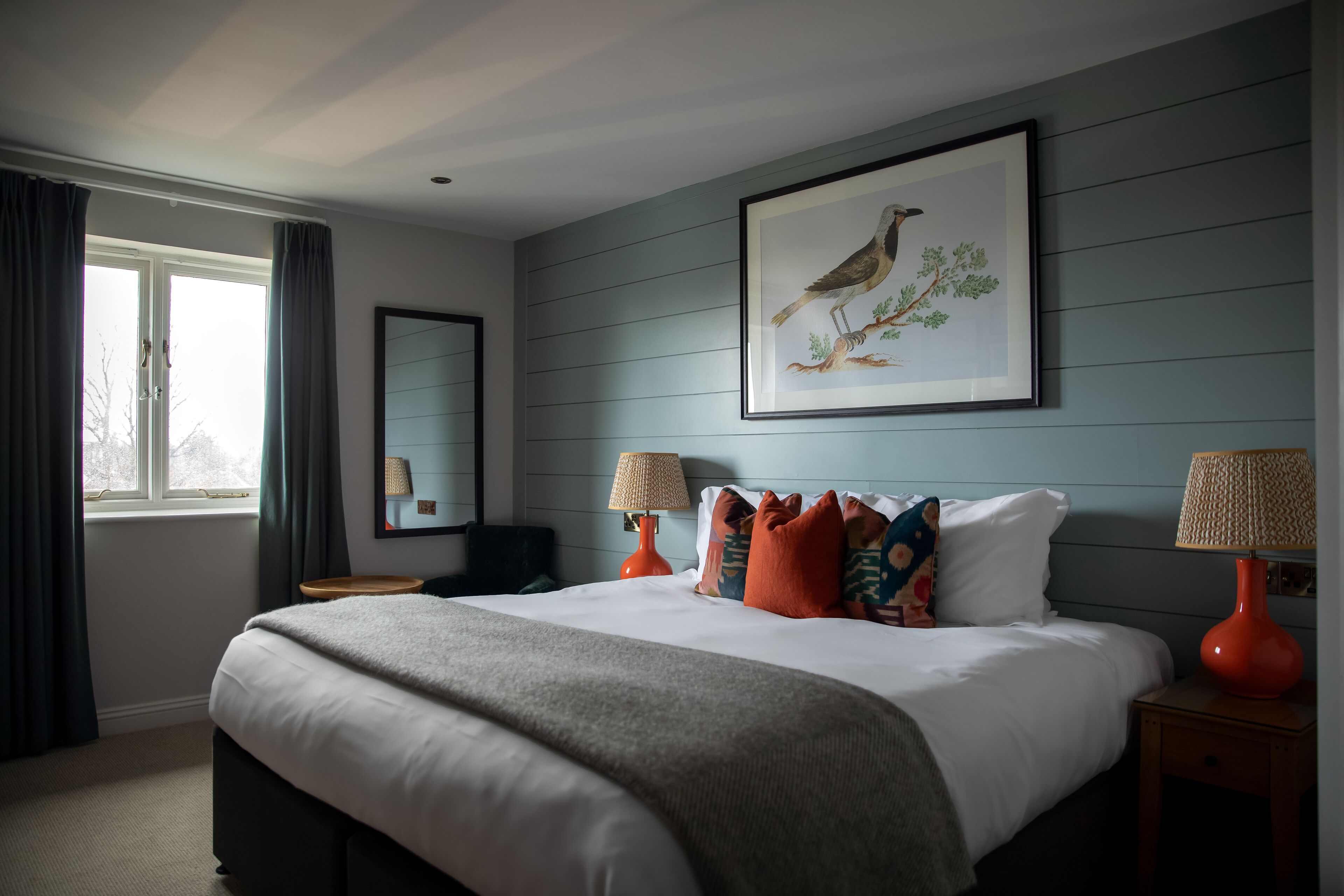 A bedroom with a bed, two bedside lamps, a framed bird painting, and a window with curtains.