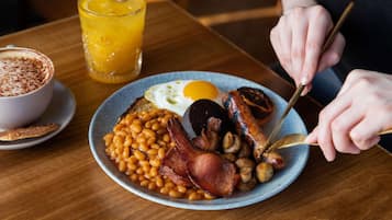 Une assiette de petit-déjeuner avec des haricots, du bacon, des champignons et un œuf au plat, un verre de jus d’orange et une tasse de café, le tout posé sur une table en bois.