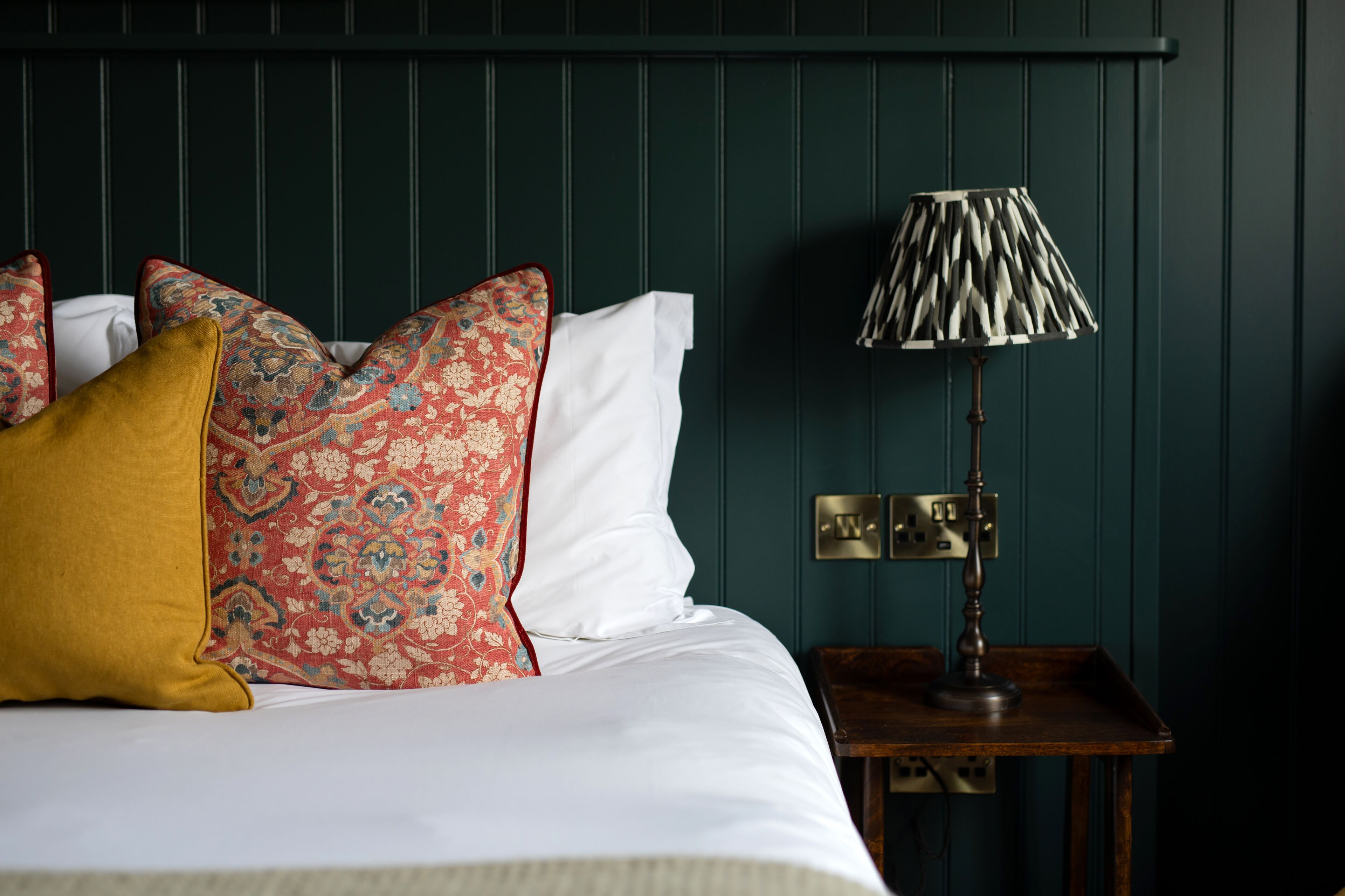 A neatly made bed with patterned pillows, a bedside lamp, and a dark green wall.